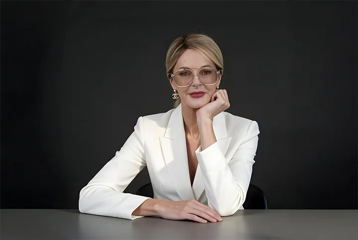 Portrait of Dr Kate Barker seated at a desk, blonde hair tied back, wearing a cream blazer and clear-framed glasses, hand resting against her cheek, photographed against a dark studio backdrop.
